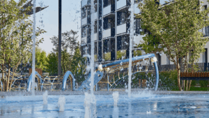 Playground with water jets spraying from the ground, modern residential building in the background and trees surrounding the area