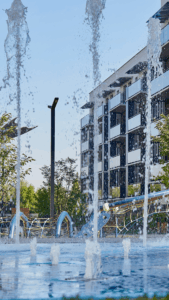 Fountain jets arch over a blue pool with a modern apartment building in the background and green trees nearby.