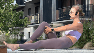 Woman in lavender athletic wear performing a boat pose on a mat outdoors, wearing a headset microphone.