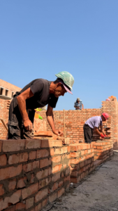 Construction worker in a green hard hat lays bricks on a low brick wall, with others building in the background under a clear sky.